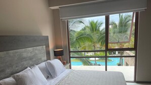 Ein Schlafzimmer mit einem großen Bett, Blick auf einen Swimmingpool und einem Balkon mit Palmen.