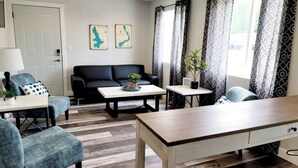 Un salon moderne avec un canapé noir, une table basse en bois, un fauteuil bleu et une table à manger. Deux plantes en pot sont posées sur des tables d’appoint, et la pièce est agrémentée de rideaux à motifs.