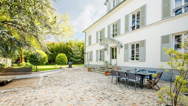 A courtyard with a stone floor, a dining area with chairs and a table, and a white building with green shutters. - 18th century residence with Jacuzzi near Paris (Mennecy)