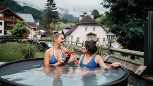 Two people in a hot tub with a scenic view of houses and mountains in the background. - Almhof Alpin Apartments & Spa (Dienten am Hochkoenig)