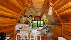 Ein Innenraum mit Holzwänden und -decke, einem zentralen Essbereich mit Hockern, einer Küchenzeile und einem großen Fenster mit Blick auf Grünflächen.