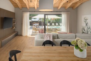 A modern living room with a wooden ceiling, a flat-screen TV mounted on a wooden panel, a grey sofa, a dining table with chairs, and a view of an outdoor pool area through glass doors. - Luxury Villa Casa Maria with pool (Tar)