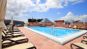 Una piscina sul tetto con lettini e ombrelloni, una struttura a cupola in vetro e un cielo azzurro limpido.