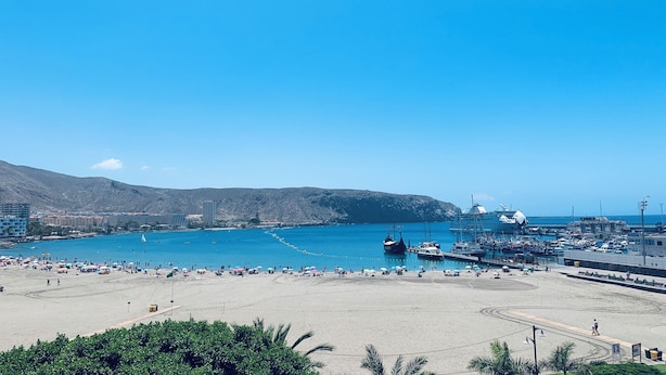 Una spiaggia costiera con un porto turistico, una riva sabbiosa e montagne sullo sfondo.