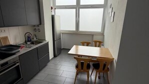 Une cuisine de taille réduite avec des meubles de couleur sombre, une table à manger avec des chaises en bois, une fenêtre à vitrage dépoli et un sol carrelé.