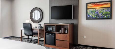 A hotel room with a bed, a desk with a chair, a flat-screen TV, a microwave, a mini-fridge, a mirror, and a painting of a field.