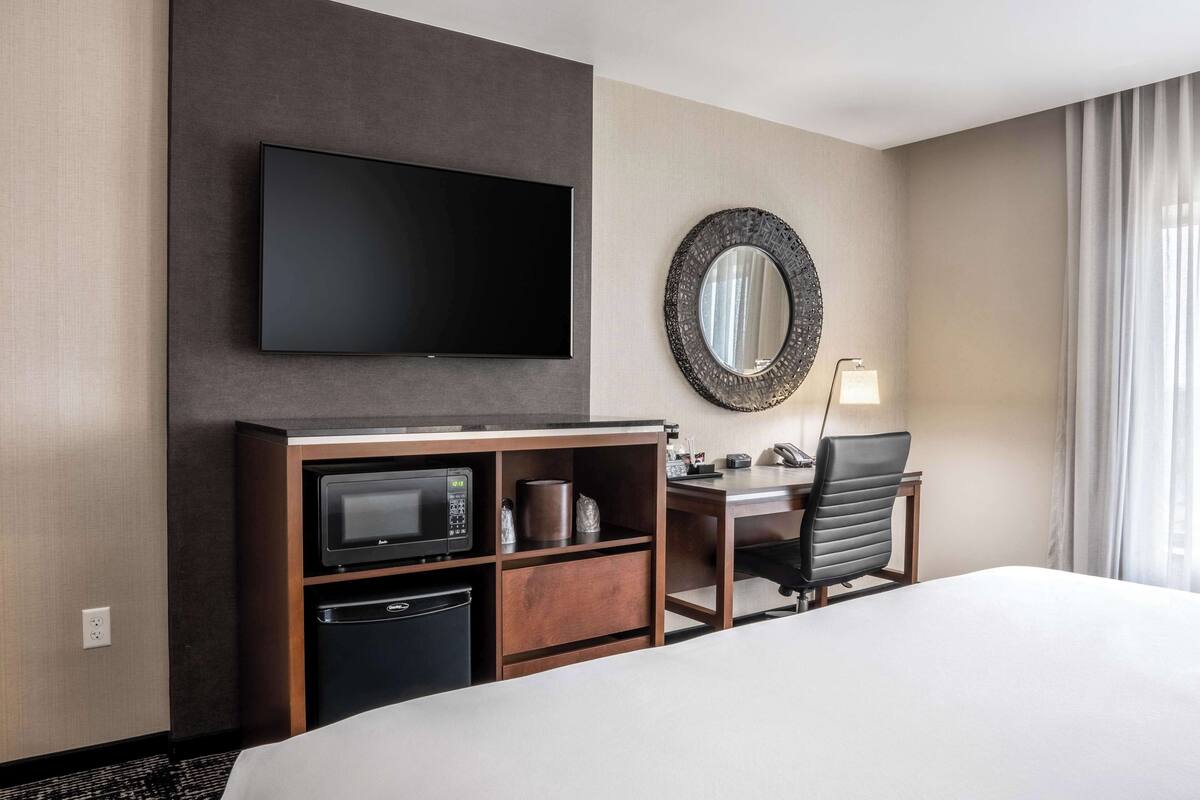 Quarto de hotel com cama, secretária com cadeira, televisão de tela plana, micro-ondas, cafeteira e um espelho grande.