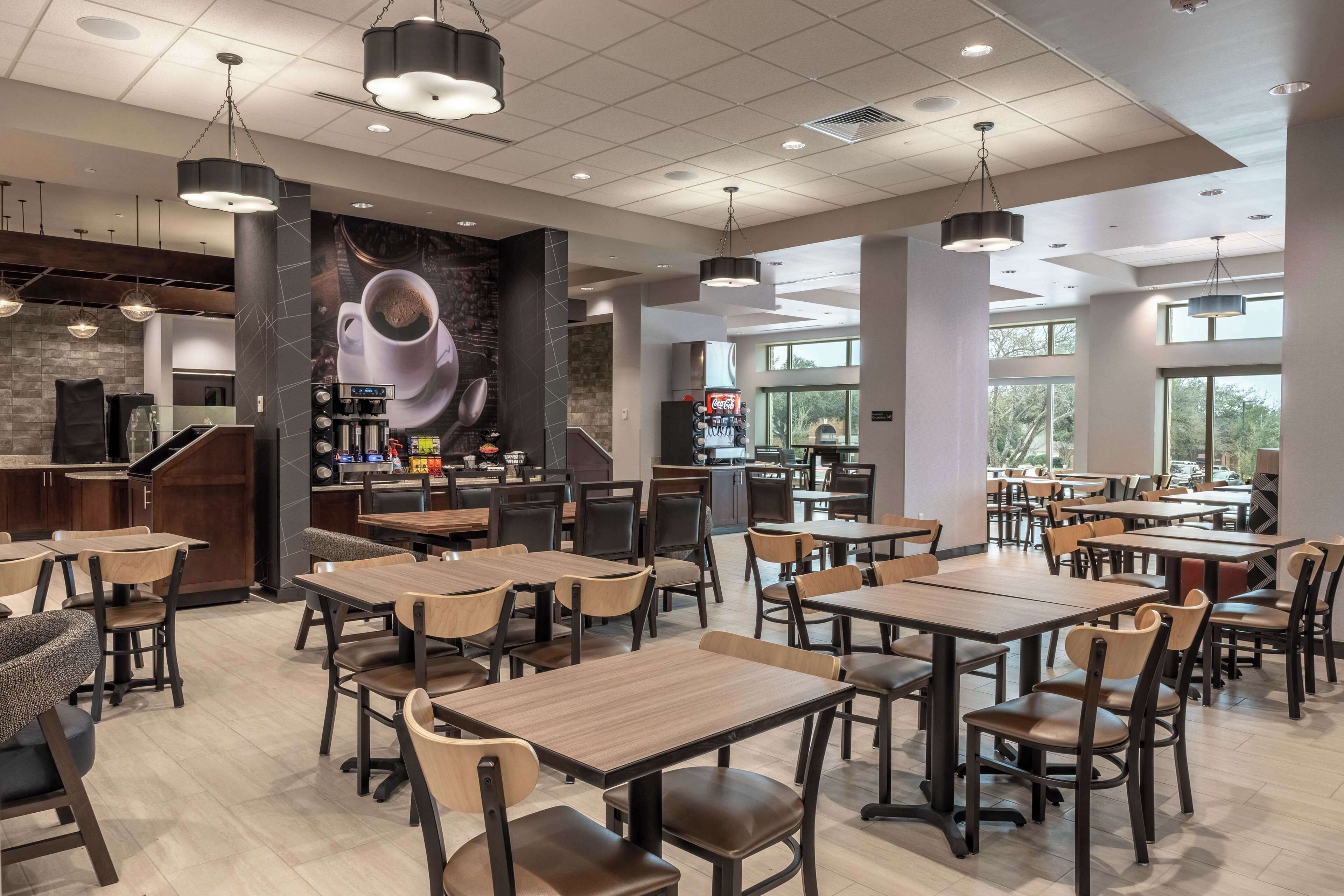 A modern cafe with multiple tables and chairs, a coffee bar, and large windows allowing natural light.
