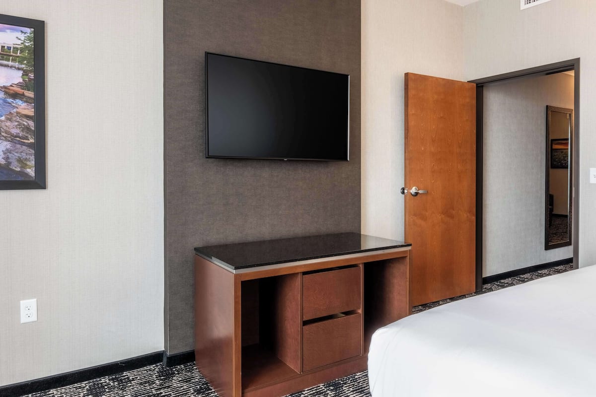 Quarto de hotel com televisão de tela plana na parede, escrivaninha de madeira com gavetas e cama com roupa de cama branca.