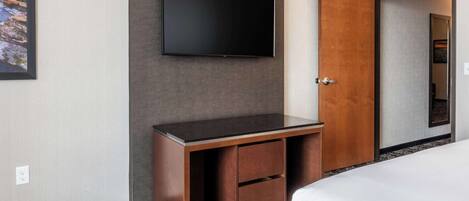 Habitación de hotel con televisión de pantalla plana montada en la pared, un escritorio de madera con cajones y una cama con ropa blanca.