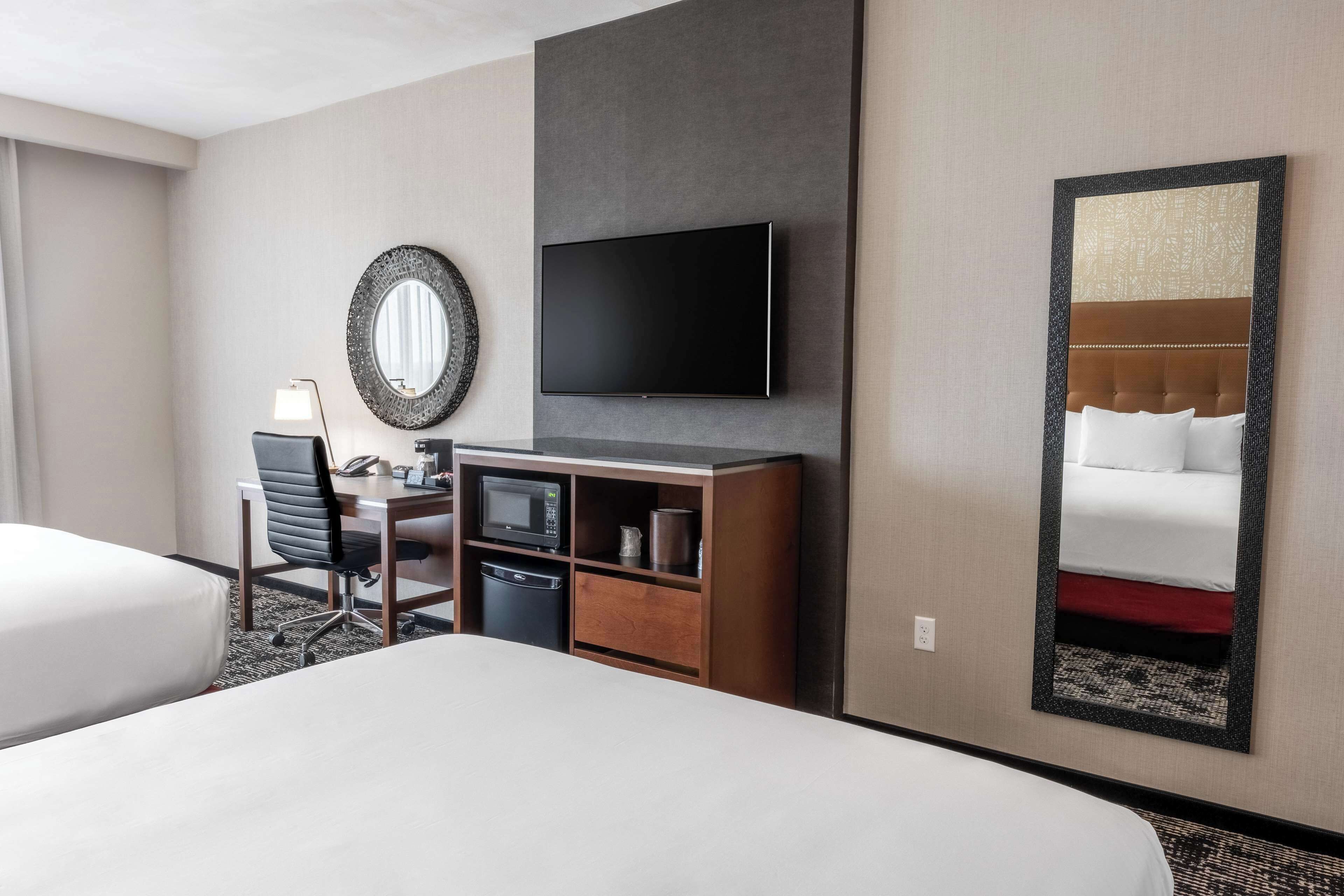 Ein Hotelzimmer mit einem Bett, einem Schreibtisch mit Stuhl, einem Flachbildfernseher, einem Spiegel und einem an der Wand befestigten Fernseher.
