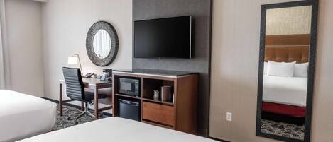 A hotel room with a bed, a desk with a chair, a flat-screen TV, a mirror, and a wall-mounted television.