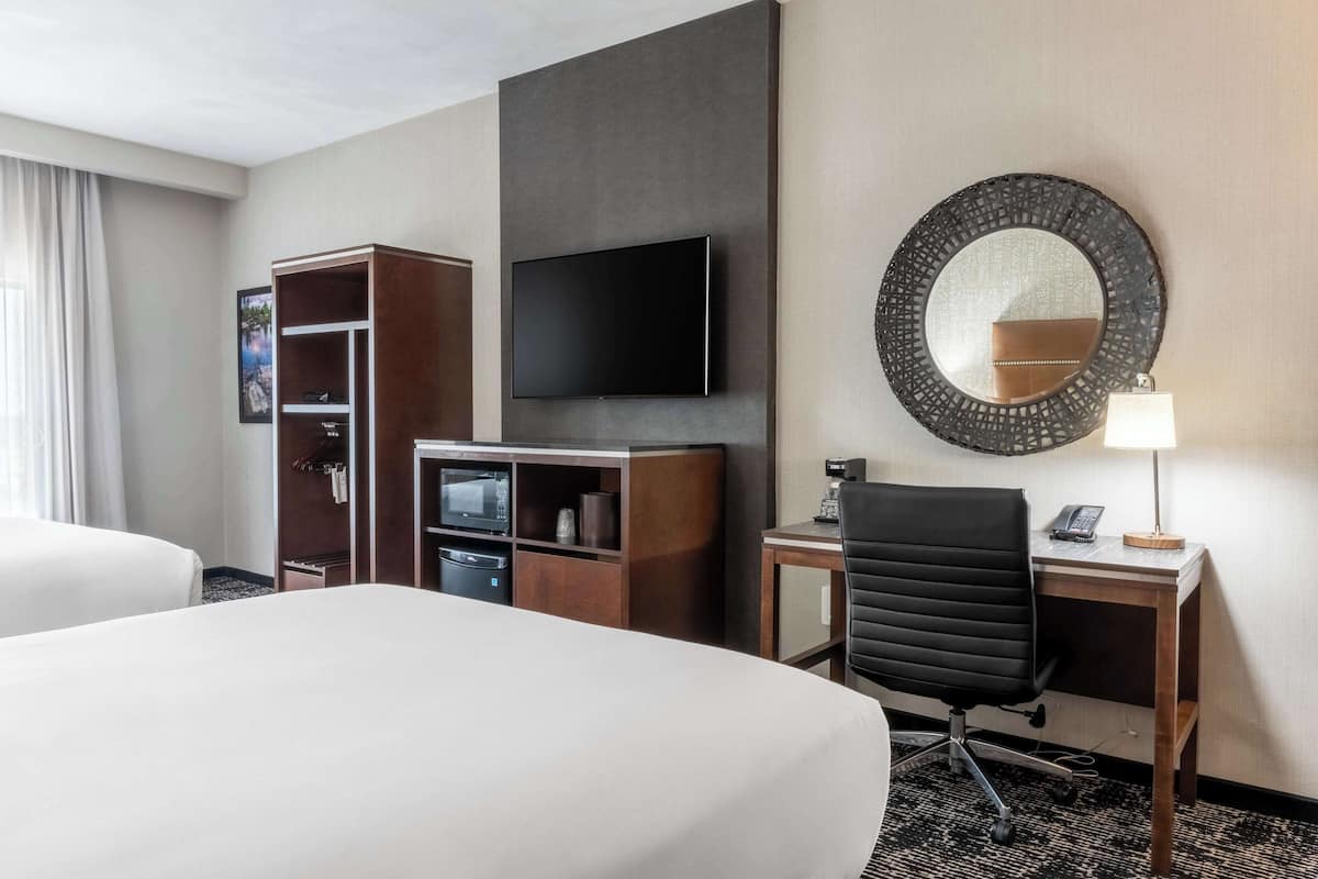Quarto de hotel com cama, escrivaninha, cadeira, televisão, espelho e guarda-roupa.
