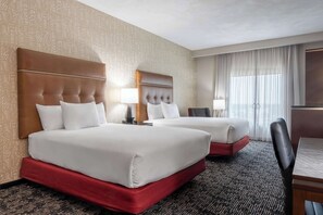 Two beds with tufted headboards and matching pillows sit on a red base, surrounded by a desk, chair, lamp, and television.