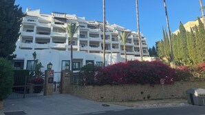 A multi-story hotel with balconies, palm trees, and a stone wall with pink flowers.