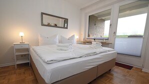 Uma cama bem arrumada com roupa de cama branca, uma mesinha de cabeceira com abajur, um espelho na parede e uma janela com vista para o exterior.