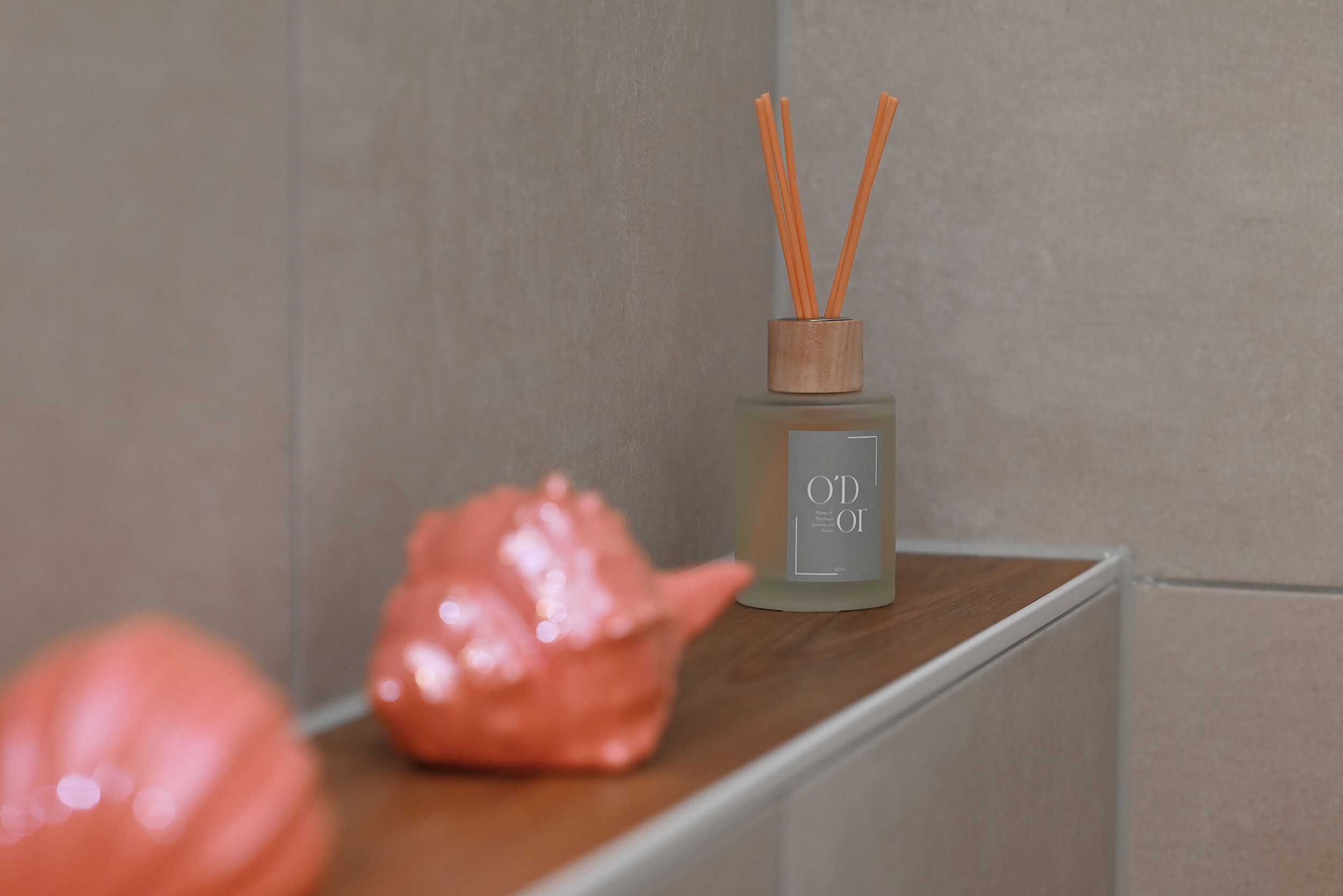 A bathroom with a beige tiled wall, a wooden shelf, and a glass bottle with orange reed sticks.