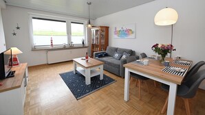 Uma sala de estar moderna com piso de madeira, um sofá cinza, uma mesa de jantar com cadeiras, um abajur e uma janela com vista.