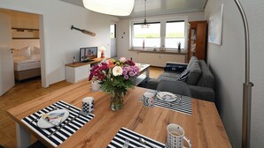 Quarto moderno e bem iluminado com uma mesa de jantar de madeira posta para dois, um sofá e vista para o exterior através de grandes janelas.