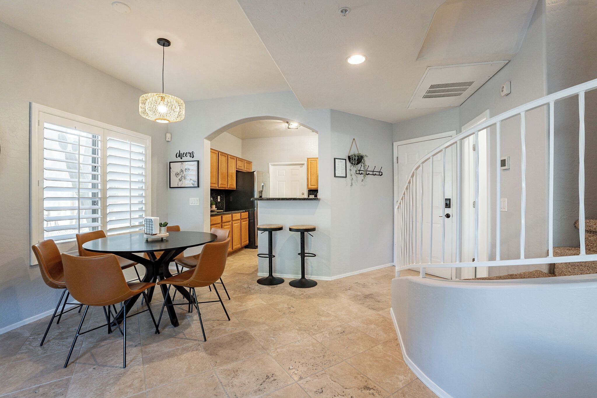 A dining area with a round table and chairs, a kitchen in the background, and a staircase with a railing.