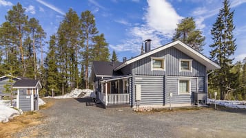 A log cabin with a porch and a small attached structure, surrounded by a gravel driveway and snow patches.