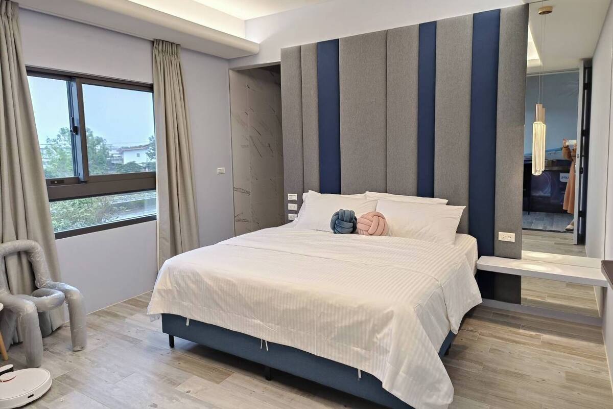 Ein modernes Schlafzimmer mit einem großen Bett, einem Sessel und Blick ins Freie durch ein Fenster.