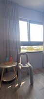 Una habitación con vista a un edificio a través de una ventana, una silla, una pequeña mesa redonda con un jarrón y una vela, y una cortina.
