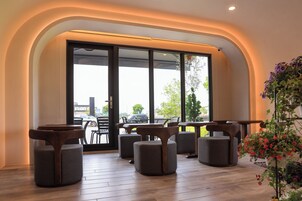 A modern interior with a seating area featuring round tables and cushioned chairs, large windows with a view of the outside, and a potted plant with flowers.