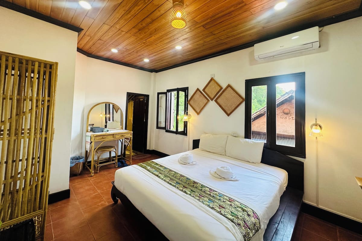 A hotel room with a wooden ceiling, a bed with white linens, a desk with a mirror, and a window with a view of greenery.