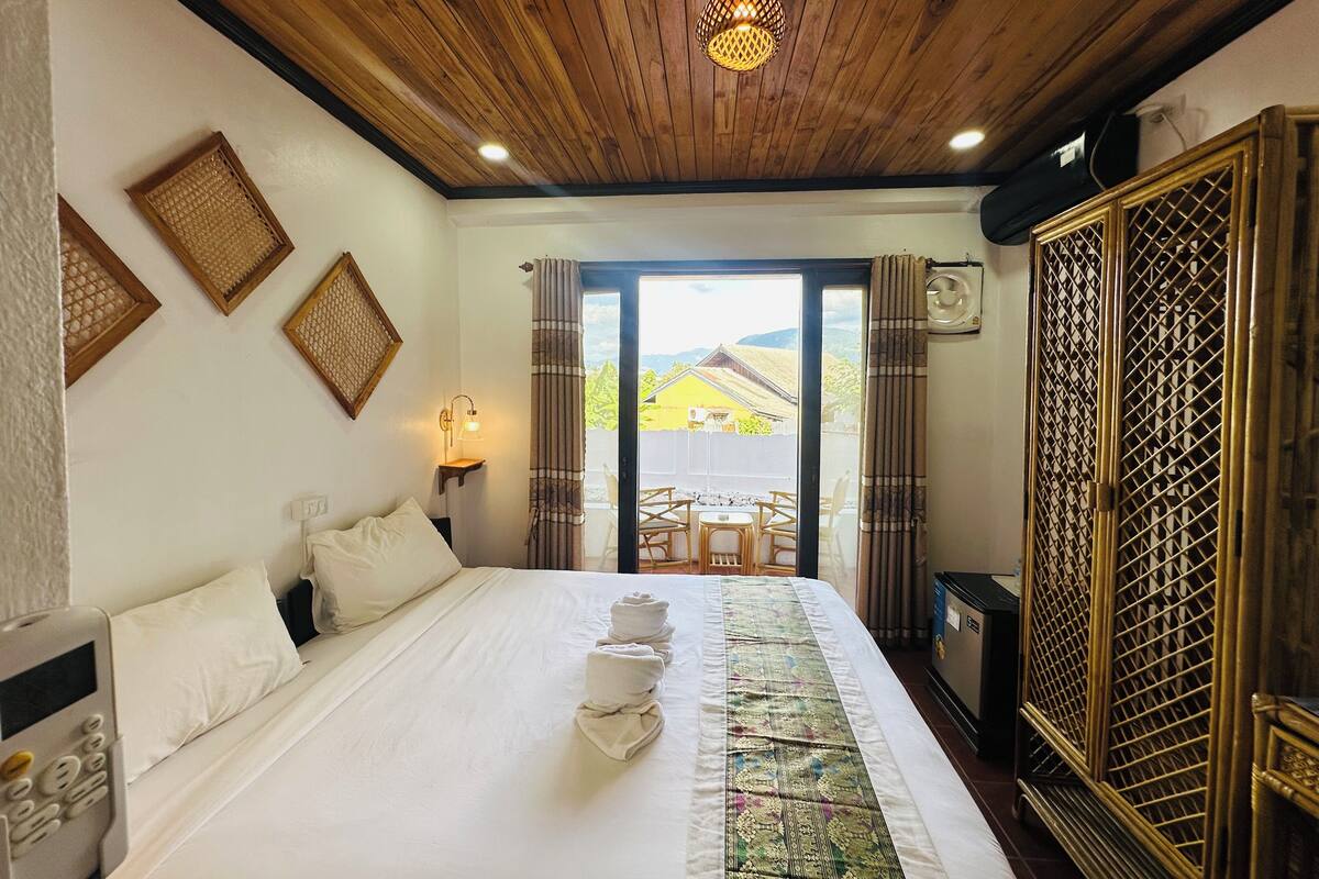 A hotel room with a wooden ceiling, a bed with white linens, a bedside table with a lamp, and a view of the outside through a sliding door.