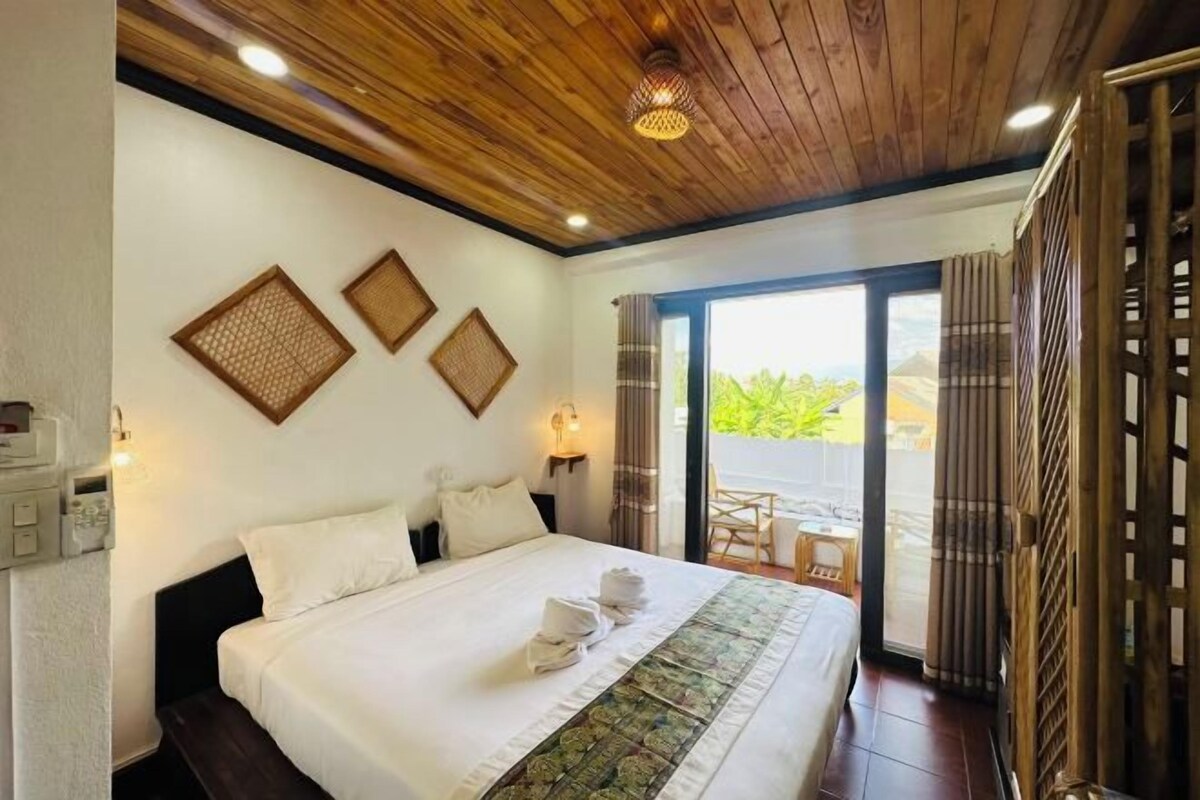 A hotel room with a wooden ceiling, a bed with white linens, a view of greenery through the glass door, and a small table with chairs outside.
