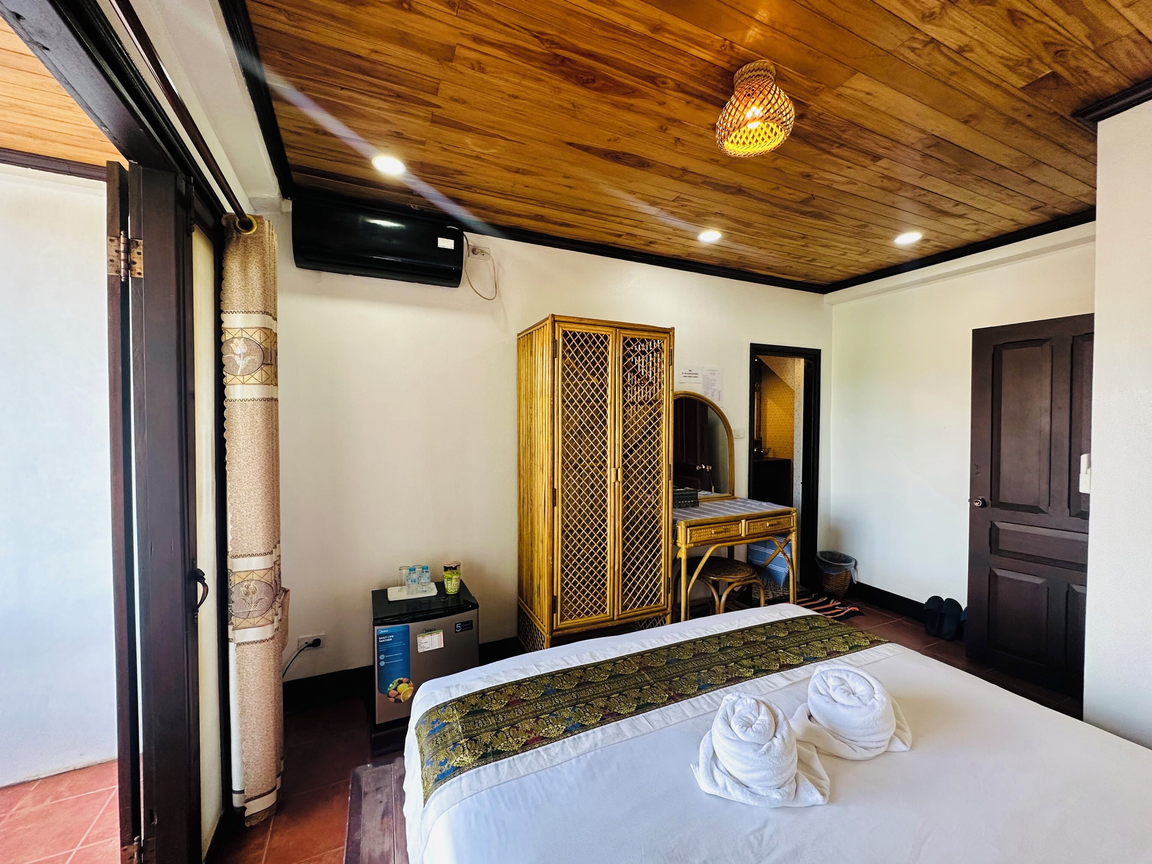 Une chambre d’hôtel avec un plafond en bois, un lit avec du linge de maison blanc et une tête de lit décorative, une armoire, un bureau avec une chaise, un miroir et un téléviseur fixé au mur.