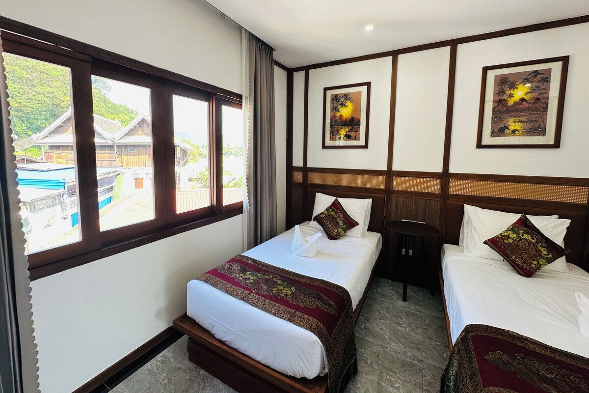 A hotel room with two beds, a window with a view of buildings, and two framed pictures on the wall.