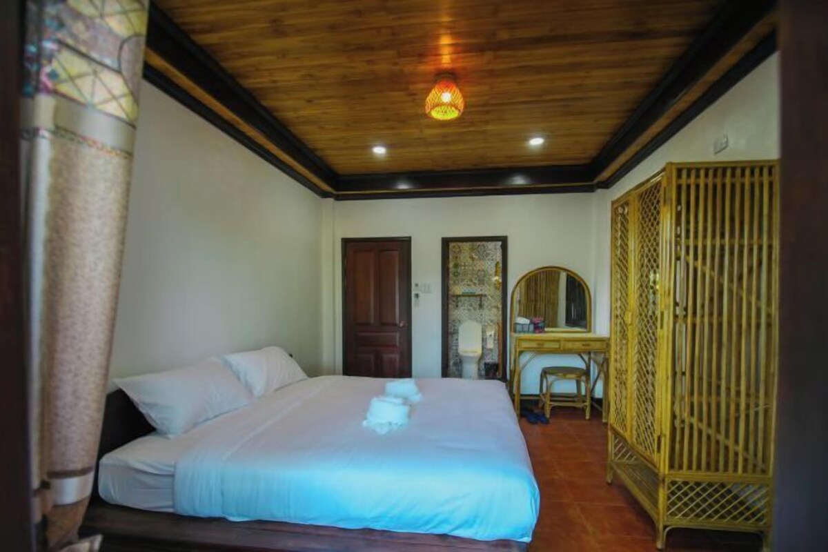 A bedroom with a wooden ceiling, a bed with white linens, a desk with a chair, and a wardrobe.
