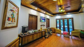 A room with a wooden ceiling, a television mounted on the wall, a motorcycle, wicker furniture, and a window with a view of the outside. - Kosy Villa (Luang Prabang)