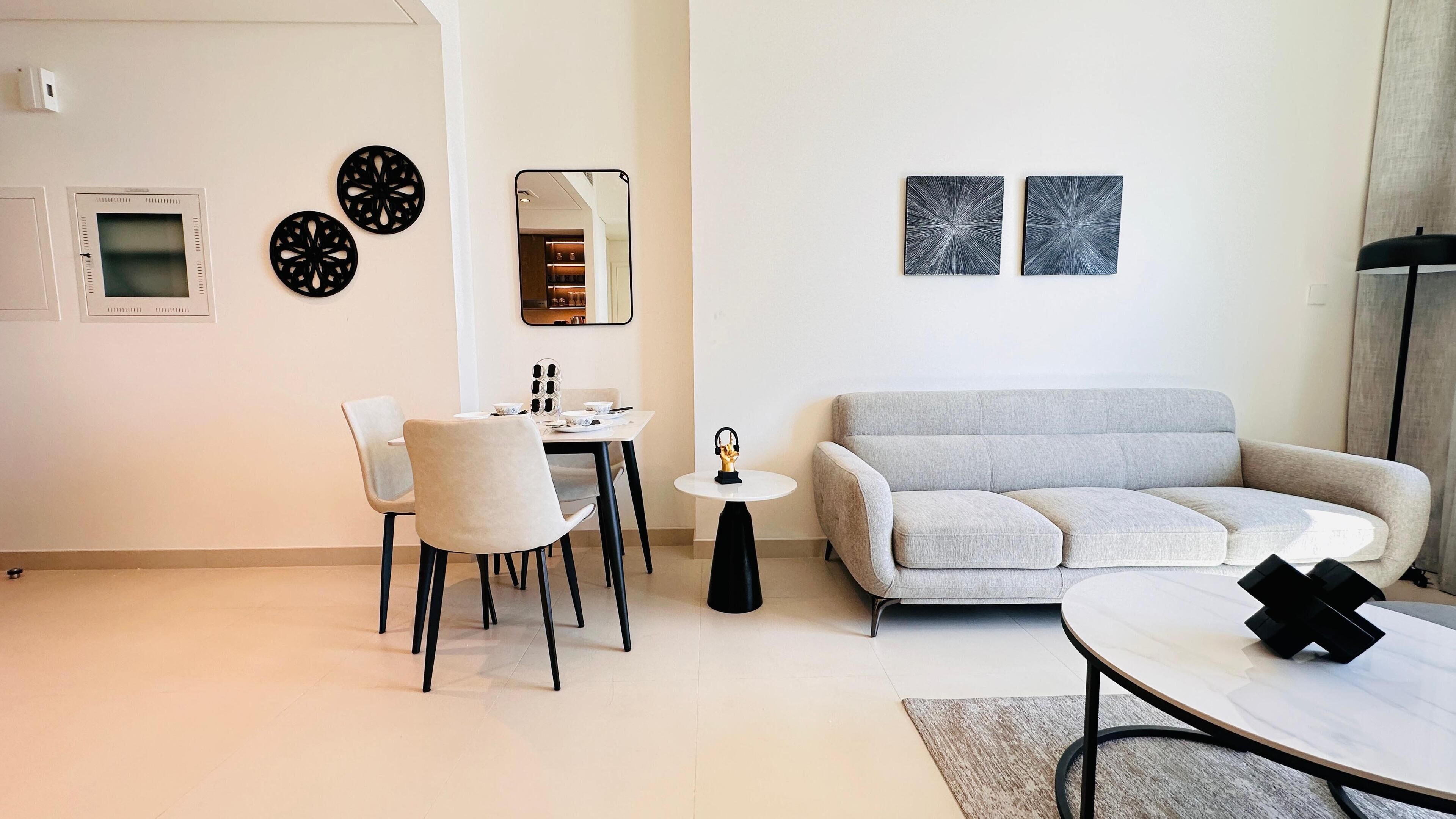 A modern living room with a grey sofa, a dining table with chairs, a small round table with a decorative item, and two abstract wall art pieces.