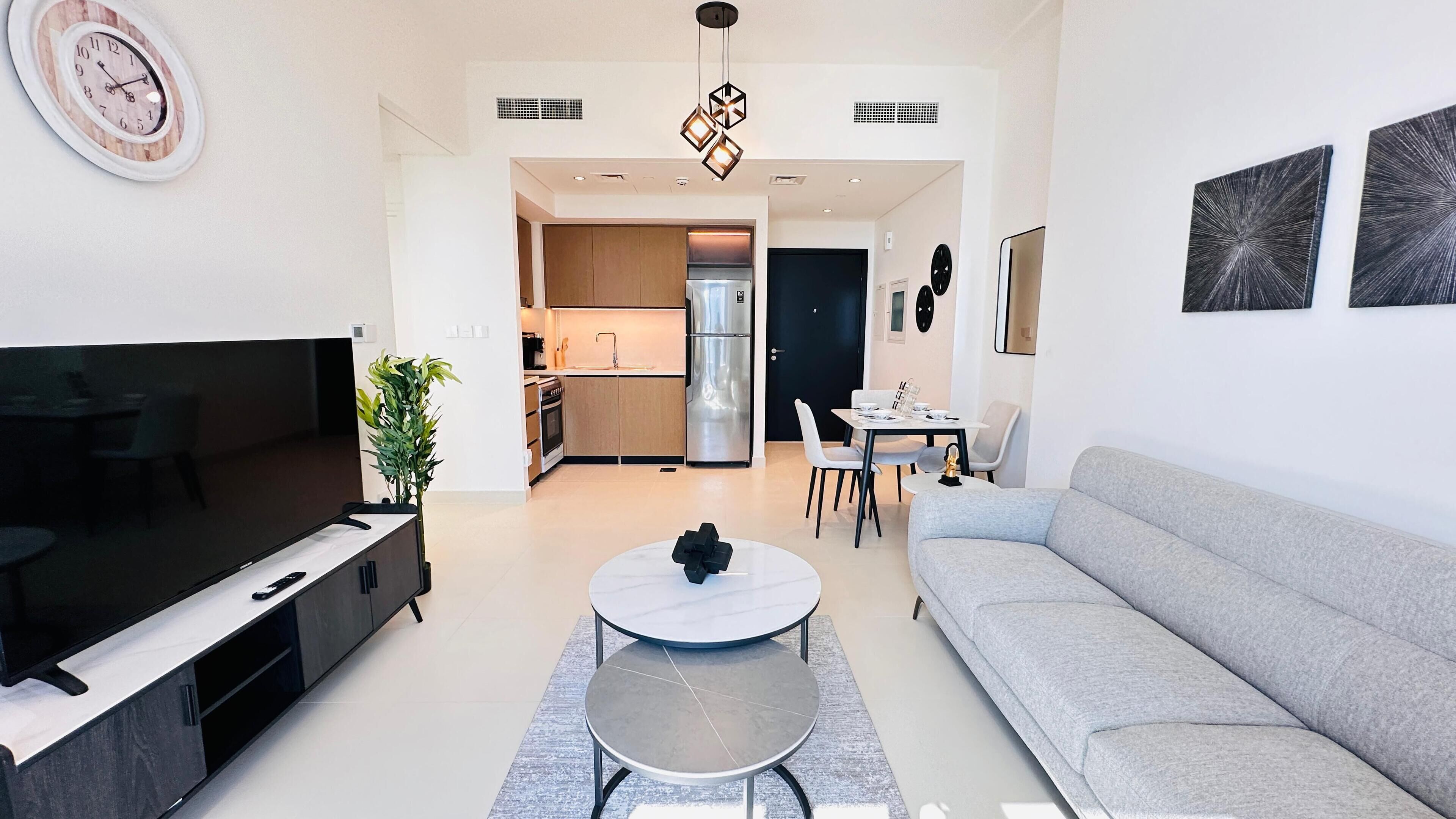 A modern living room with a flat-screen TV, a grey sofa, a round coffee table, and a dining area with a white table and chairs.