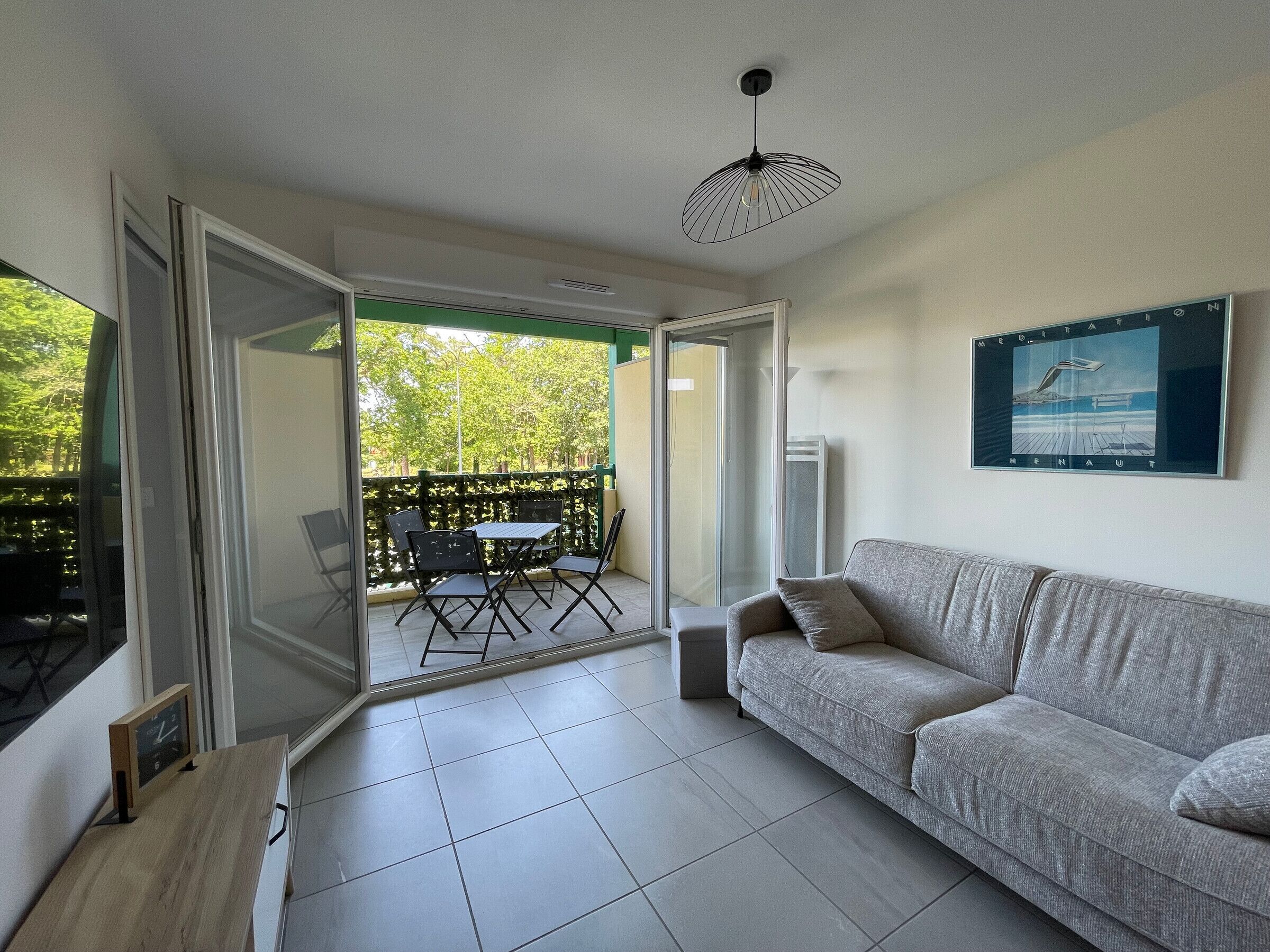 A modern living room with a grey sofa, a small table, and a chair. There is a balcony with a table and chairs outside.