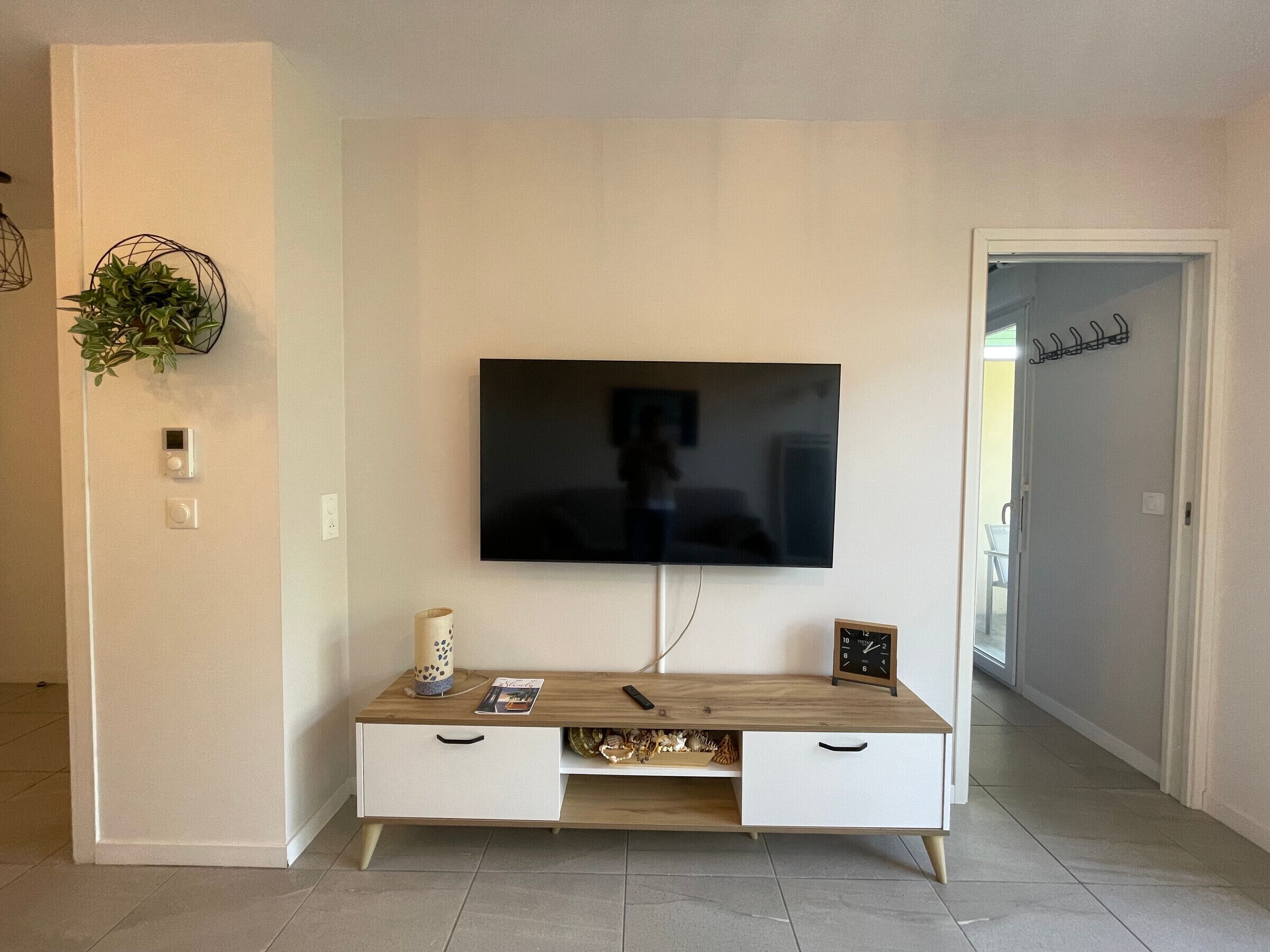 A modern living room with a flat-screen TV mounted on the wall, a wooden coffee table with a plant, and a white cabinet with drawers.