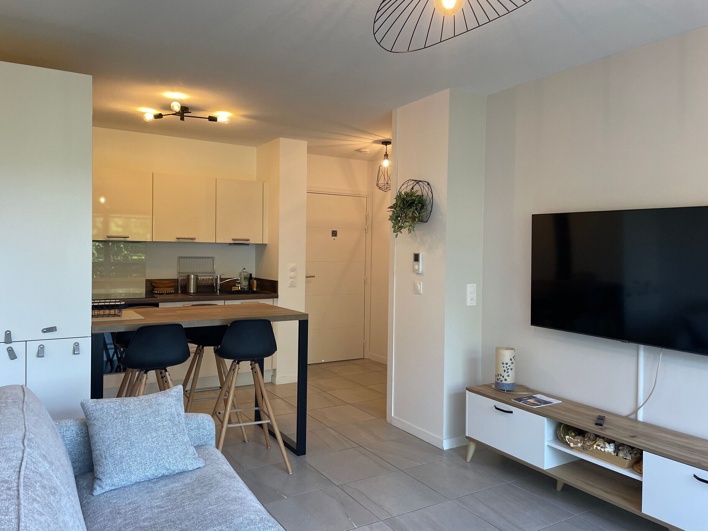 Uma sala de estar moderna com um sofá cinzento, uma TV na parede e uma cozinha ao fundo.