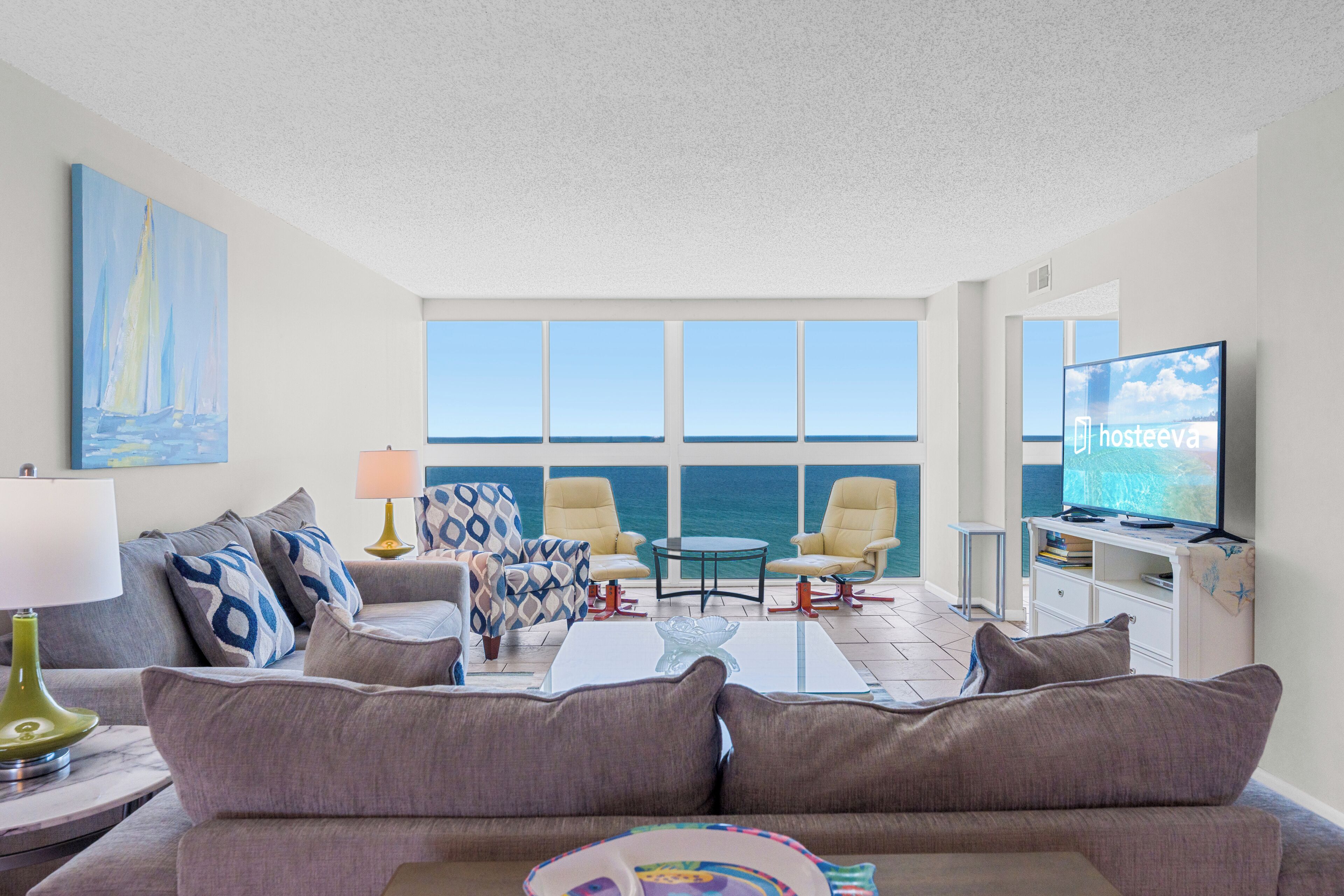 A living room with a sofa, armchairs, a coffee table, and a TV. There are large windows with a view of the sea.