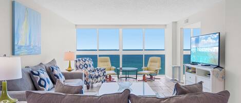 A living room with a sofa, armchairs, a coffee table, and a TV. There are large windows with a view of the sea.