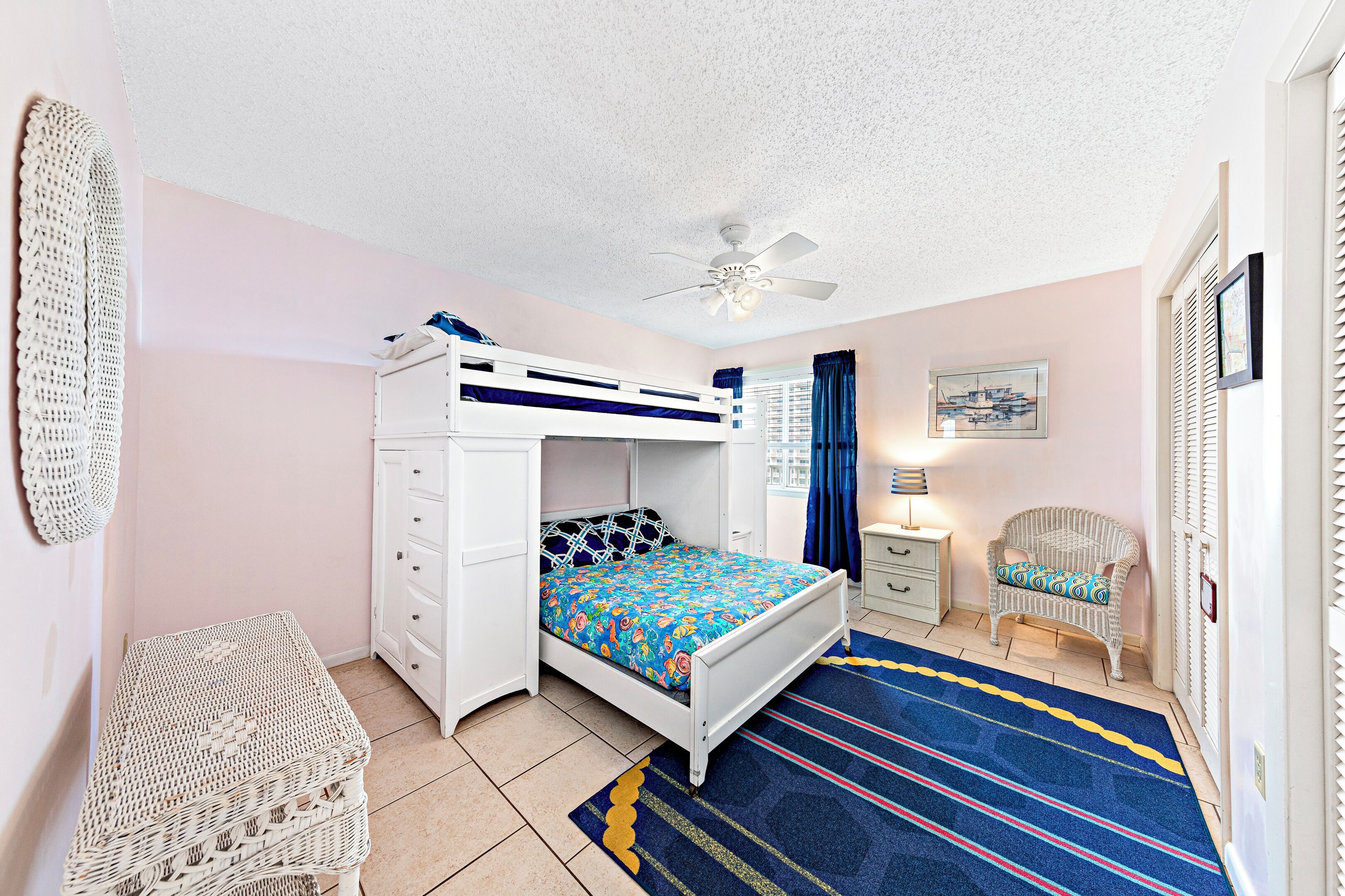 A bedroom with a bunk bed, a white dresser, a wicker chair, a small table, and a ceiling fan.