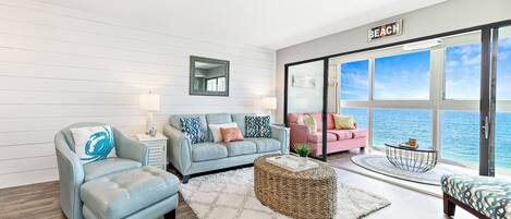 A modern living room with a light blue sofa, a matching armchair, and a pink sofa. There is a large window with a view of the ocean.