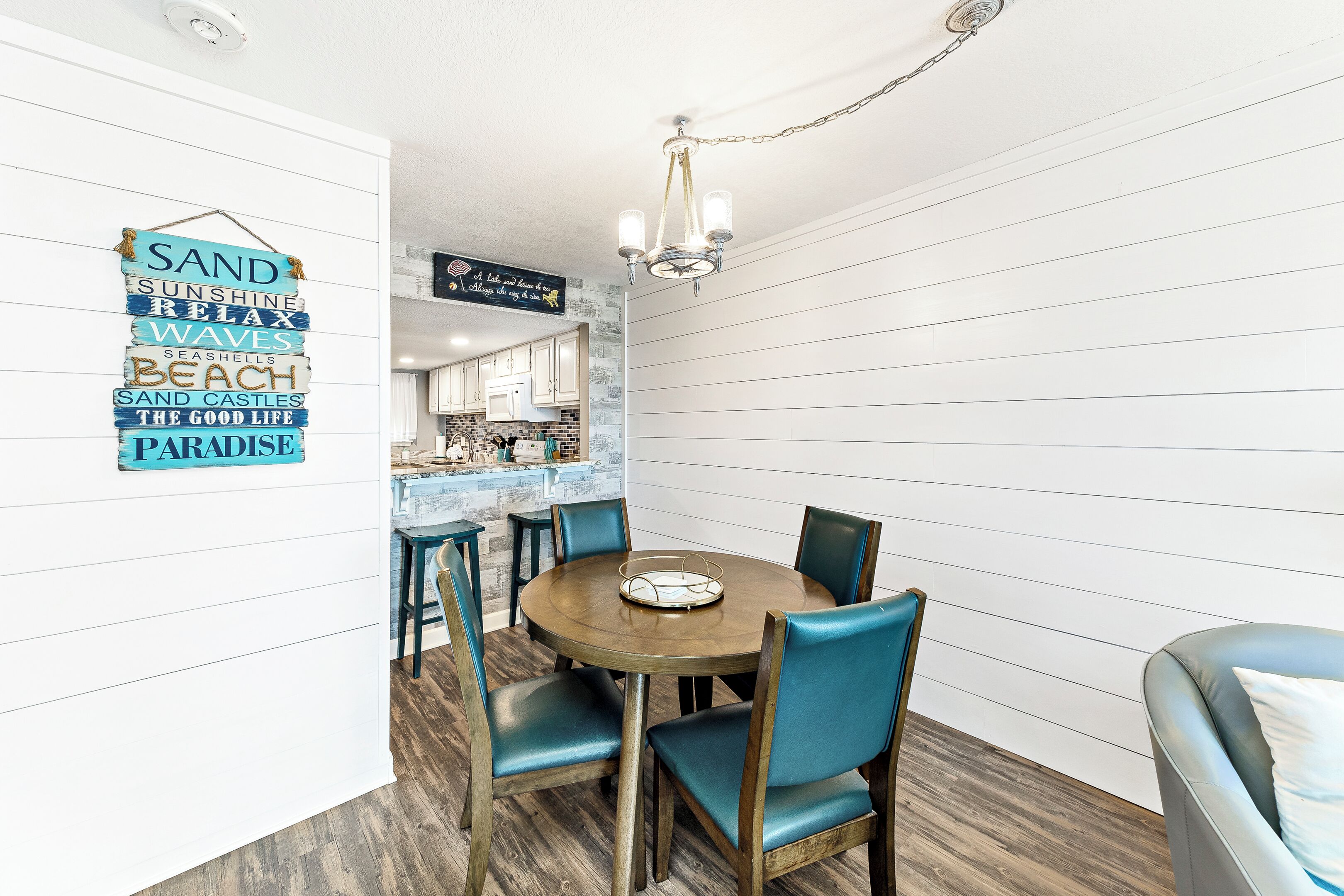 A dining area with a round wooden table and blue upholstered chairs. A hanging sign with words like "SAND," "SUNSHINE," and "BEACH" is visible.