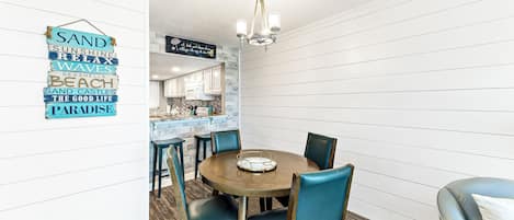 A dining area with a round wooden table and blue upholstered chairs. A hanging sign with words like "SAND," "SUNSHINE," and "BEACH" is visible.