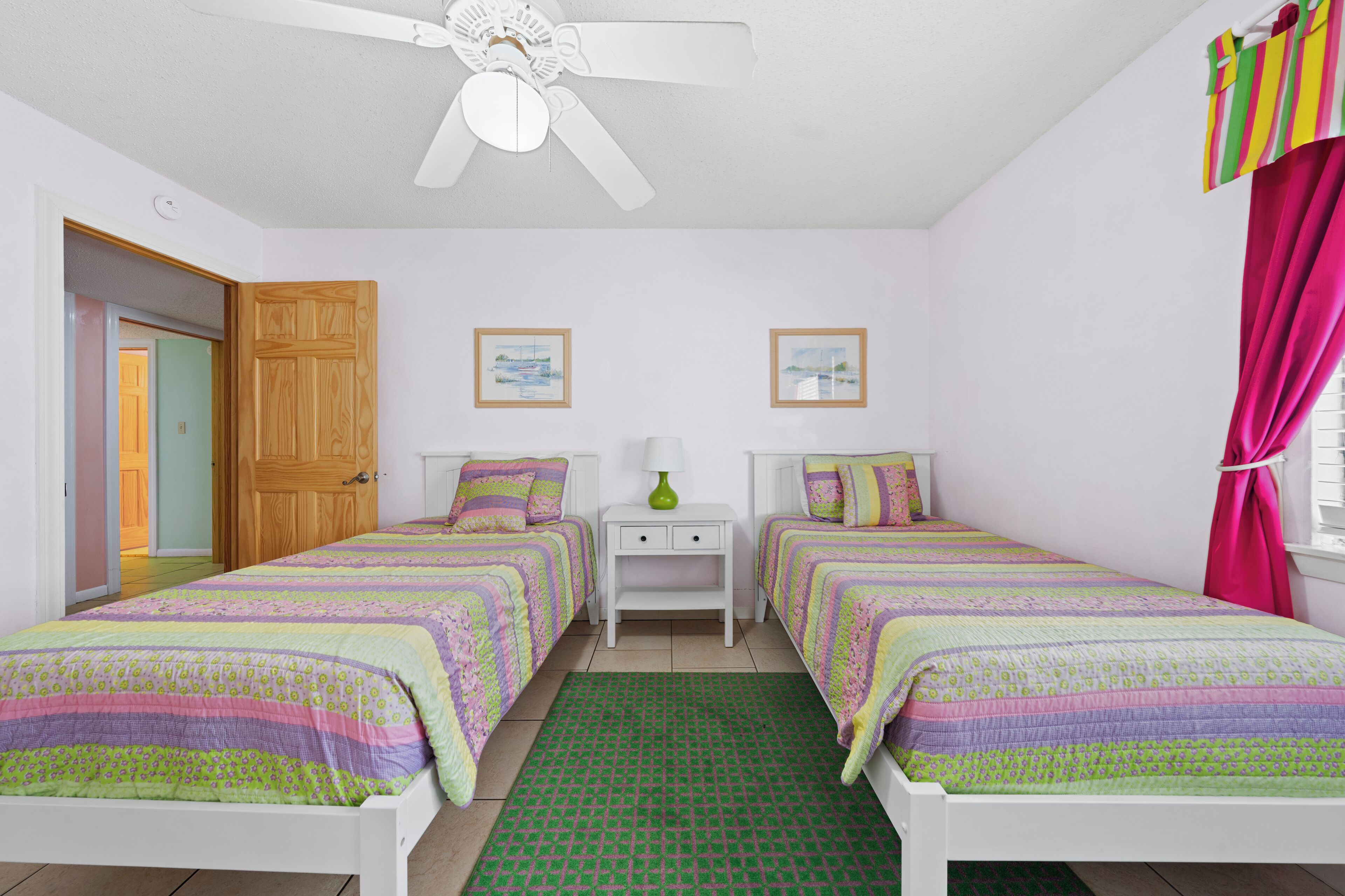 Un dormitorio con dos camas individuales, un ventilador de techo, una mesita de noche con lámpara y una alfombra verde.