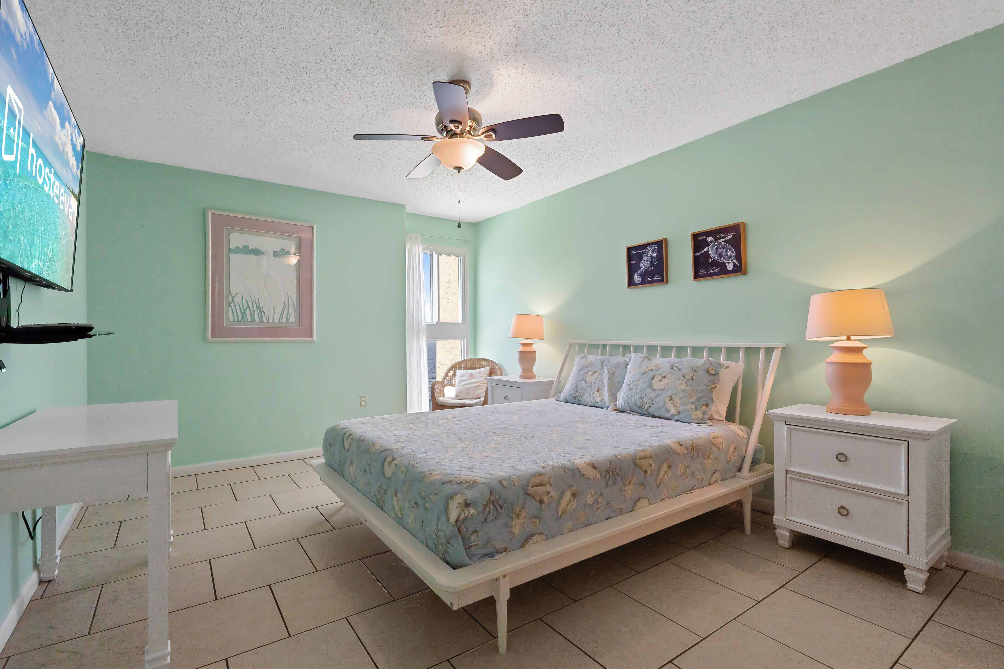 A bedroom with a bed, nightstands, a ceiling fan, a TV, and artwork on the walls.