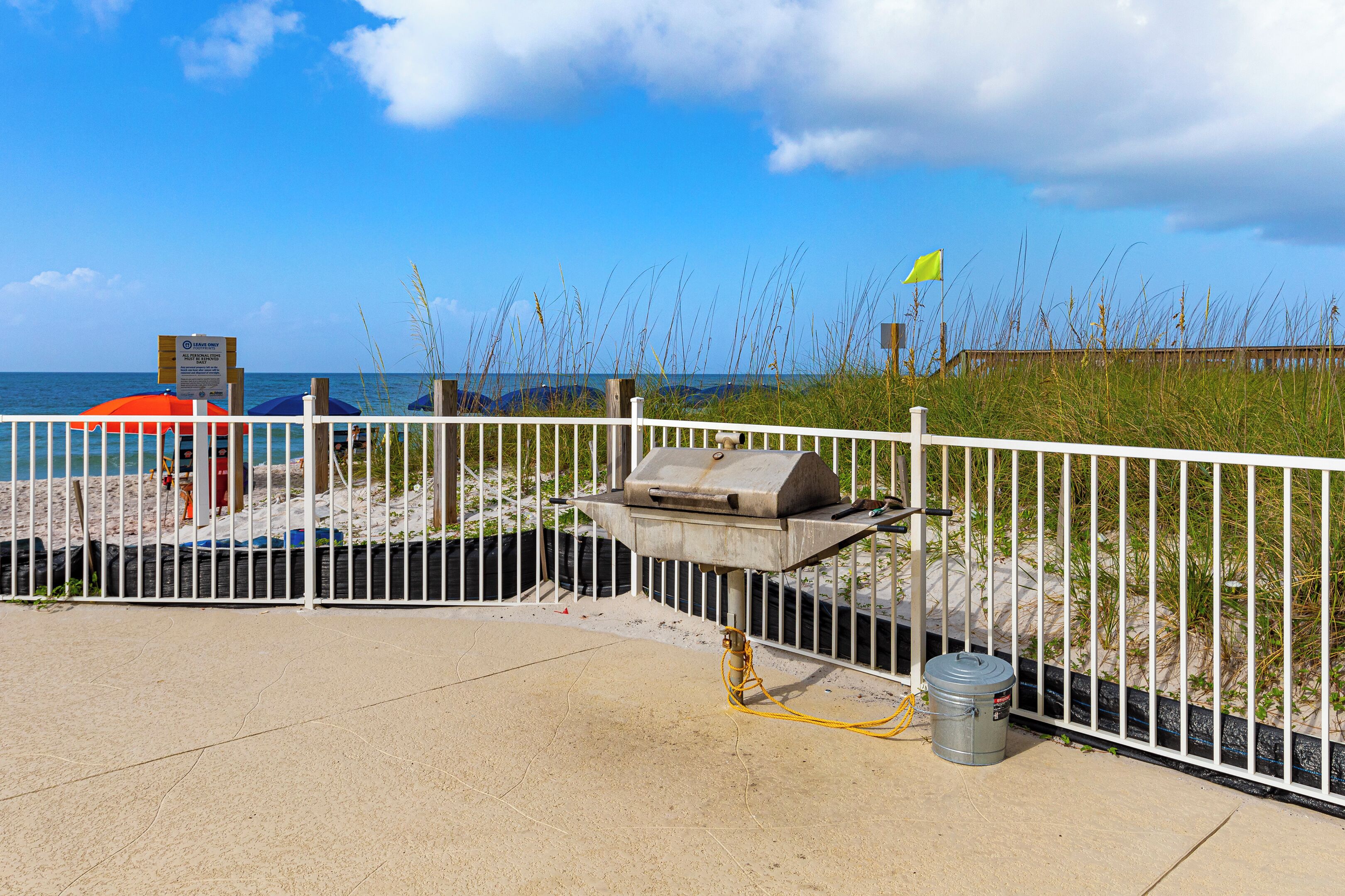 A beachside barbecue area with a grill, a trash can, and a white fence.