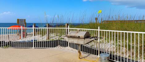 A beachside barbecue area with a grill, a trash can, and a white fence.